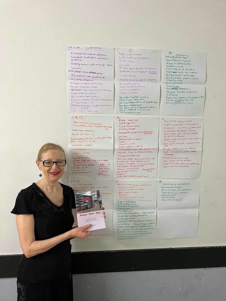 Hilaire, dressed in black and smiling, stands in front of a wall to which A3 sheets of paper covered in hand written words and phrases are attached. She is holding a copy of the found ~ flow ~ flux booklet.